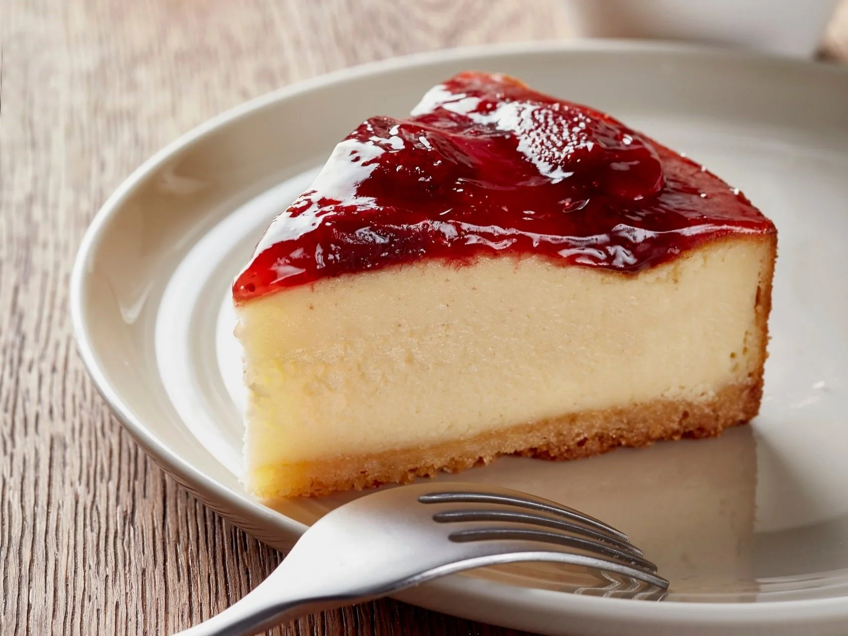 Slice of creamy cheesecake topped with red fruit glaze on a beige plate, with a fork resting beside it.